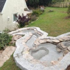 A partially constructed multi-level garden pond made of concrete and stone sits on a grassy slope beside a white house, showcasing property refurbishments, with plants and trees in the background.