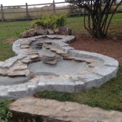 A dry stone water feature under construction by a skilled builder showcases a curved, multi-level shape with stone edging in the yard; nearby, a bare patch of soil sits beside a wooden fence and grassy field—ideal for property refurbishments.