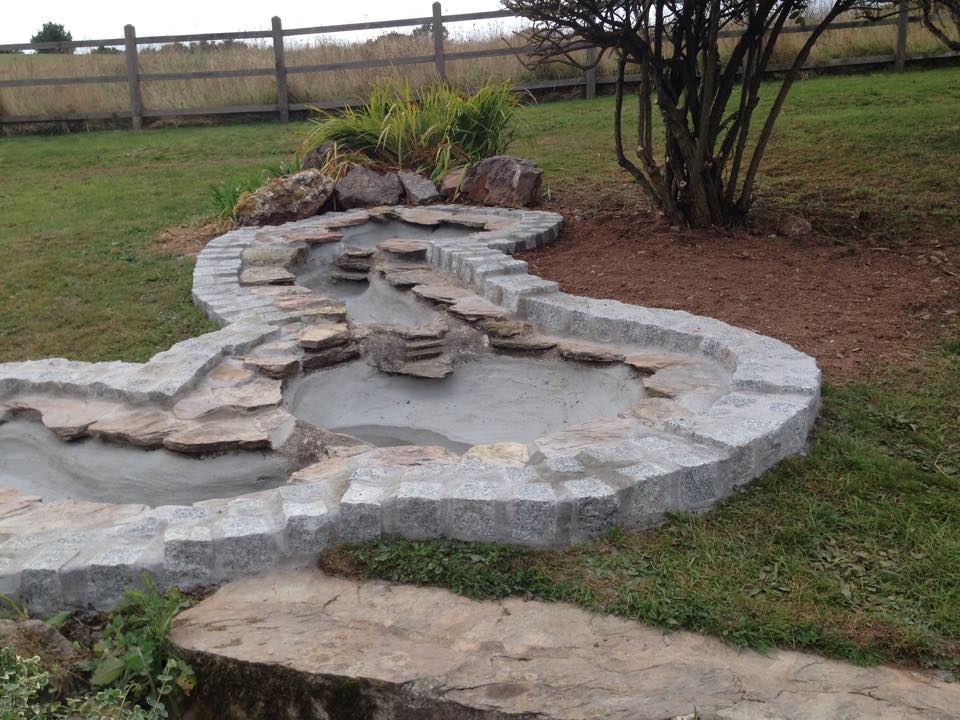 A dry stone water feature under construction by a skilled builder showcases a curved, multi-level shape with stone edging in the yard; nearby, a bare patch of soil sits beside a wooden fence and grassy field—ideal for property refurbishments.