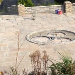A circular stone feature with three metal rings is set into a paved patio area, suggesting the craftsmanship of a skilled builder. Low stone walls, garden plants, and two plastic buckets complement the scene, with trees visible in the background.