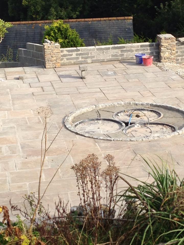 A circular stone feature with three metal rings is set into a paved patio area, suggesting the craftsmanship of a skilled builder. Low stone walls, garden plants, and two plastic buckets complement the scene, with trees visible in the background.