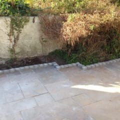 A paved patio area with a raised garden bed bordered by stone bricks. The garden bed contains overgrown, dry plants and vines climbing a concrete wall—perfect for a builder to enhance alongside new bathrooms or kitchen installations. Sunlight casts shadows on the tiles.