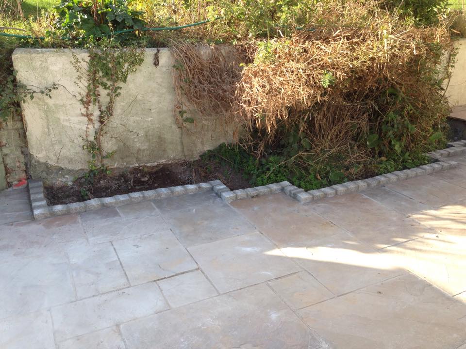 A paved patio area with a raised garden bed bordered by stone bricks. The garden bed contains overgrown, dry plants and vines climbing a concrete wall—perfect for a builder to enhance alongside new bathrooms or kitchen installations. Sunlight casts shadows on the tiles.