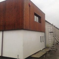 A modern two-story building with white walls below and wood paneling above, featuring several windows and a tall metal ladder—ideal for property refurbishments or kitchen installations. The sky is overcast.