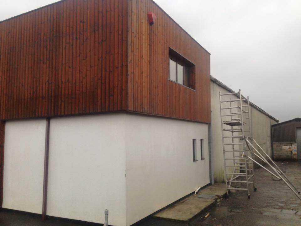 A modern two-story building with white walls below and wood paneling above, featuring several windows and a tall metal ladder—ideal for property refurbishments or kitchen installations. The sky is overcast.