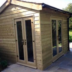 A small wooden garden shed with double glass doors and side windows, set on a stone patio and surrounded by greenery—a perfect space for garden tools or as a builder’s workshop, blending charm with practical building maintenance.