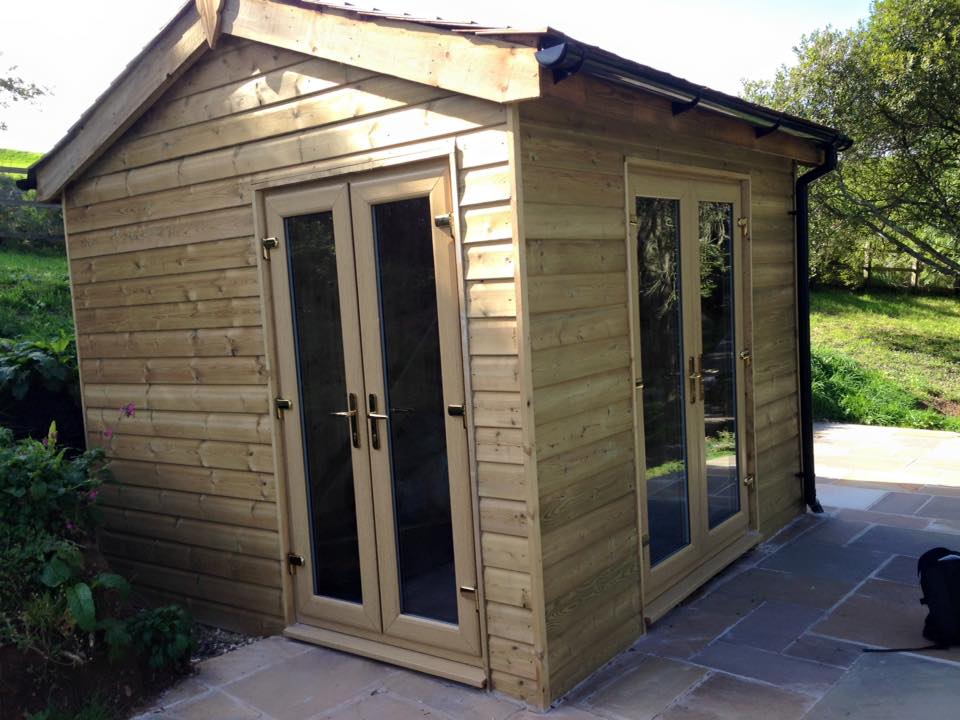 A small wooden garden shed with double glass doors and side windows, set on a stone patio and surrounded by greenery—a perfect space for garden tools or as a builder’s workshop, blending charm with practical building maintenance.