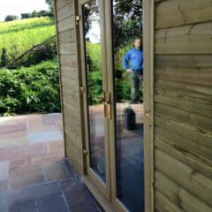 A pair of glass double doors with wooden frames are set into a timber-clad building, reflecting a man outside on a stone patio. This property showcases quality building maintenance with lush greenery and fields in the background.