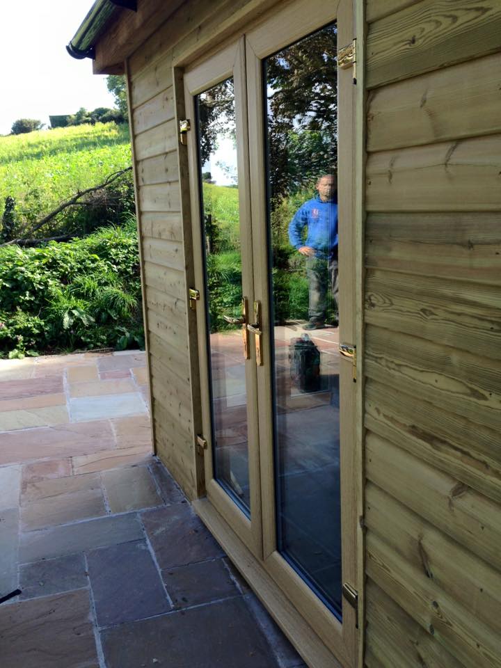 A pair of glass double doors with wooden frames are set into a timber-clad building, reflecting a man outside on a stone patio. This property showcases quality building maintenance with lush greenery and fields in the background.