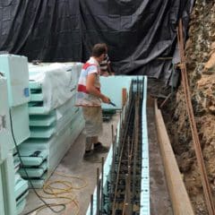 A builder in work clothes assembles insulated concrete forms (ICFs) with steel reinforcement bars on a construction site, next to a tarp-covered wall and a pile of stacked foam panels—ideal for property refurbishments.