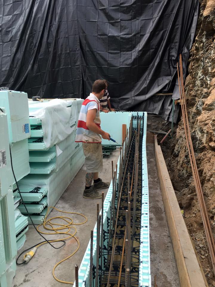 A builder in work clothes assembles insulated concrete forms (ICFs) with steel reinforcement bars on a construction site, next to a tarp-covered wall and a pile of stacked foam panels—ideal for property refurbishments.