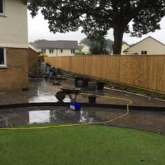 A backyard with wet stone paving, a wheelbarrow, a hose, and several black pots. A large tree and wooden fence stand near houses in the background. The damp weather sets the scene for outdoor building maintenance projects.