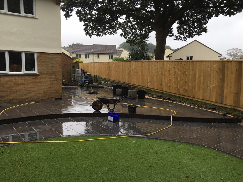 A backyard with wet stone paving, a wheelbarrow, a hose, and several black pots. A large tree and wooden fence stand near houses in the background. The damp weather sets the scene for outdoor building maintenance projects.