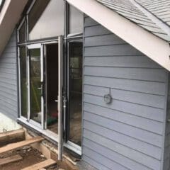 A gray house with horizontal siding, large glass doors, and exposed wooden beams on the patio area under construction. The roof has gray shingles, and the builder is focusing on kitchen installations while exterior building maintenance is ongoing.
