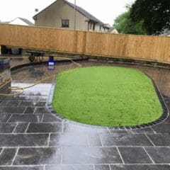 A backyard with wet black stone tiles, an oval-shaped artificial grass area in the center, a wooden fence in the background, and a yellow garden hose lying on the ground—ideal for property refurbishments or builder projects.