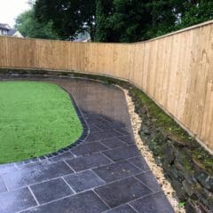 A wet, curved stone patio borders a section of artificial grass and a raised stone wall, with a tall wooden fence and trees in the background—an ideal setting for property refurbishments or new bathrooms.