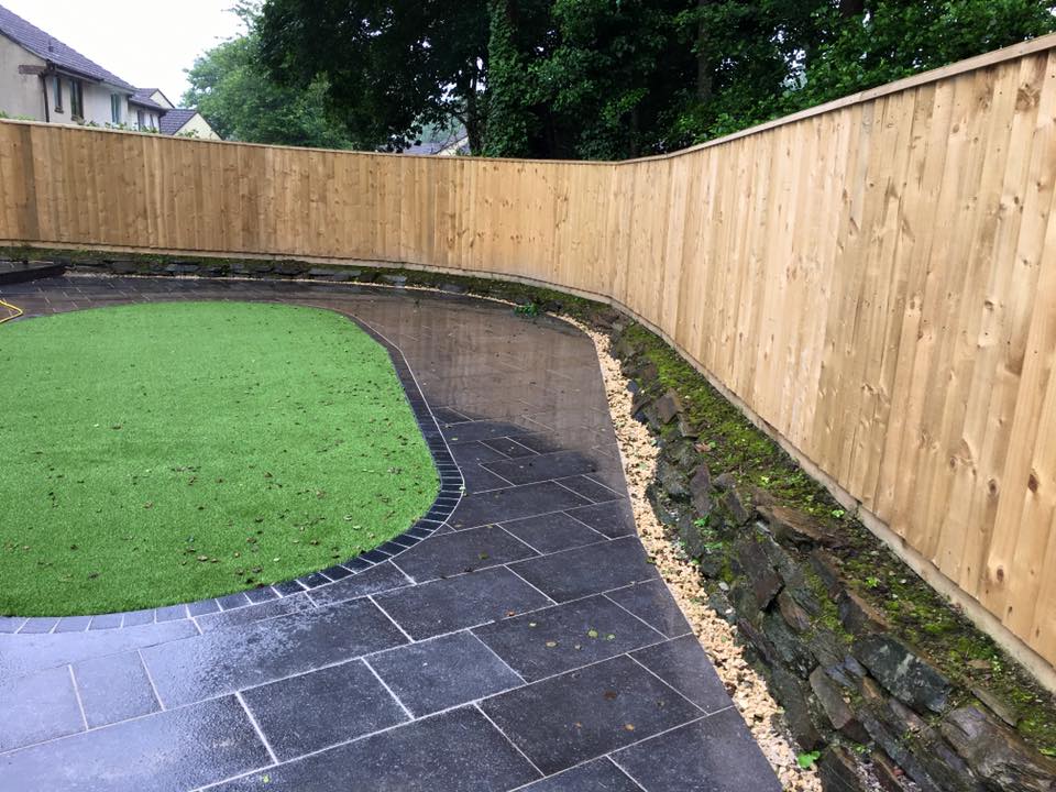 A wet, curved stone patio borders a section of artificial grass and a raised stone wall, with a tall wooden fence and trees in the background—an ideal setting for property refurbishments or new bathrooms.