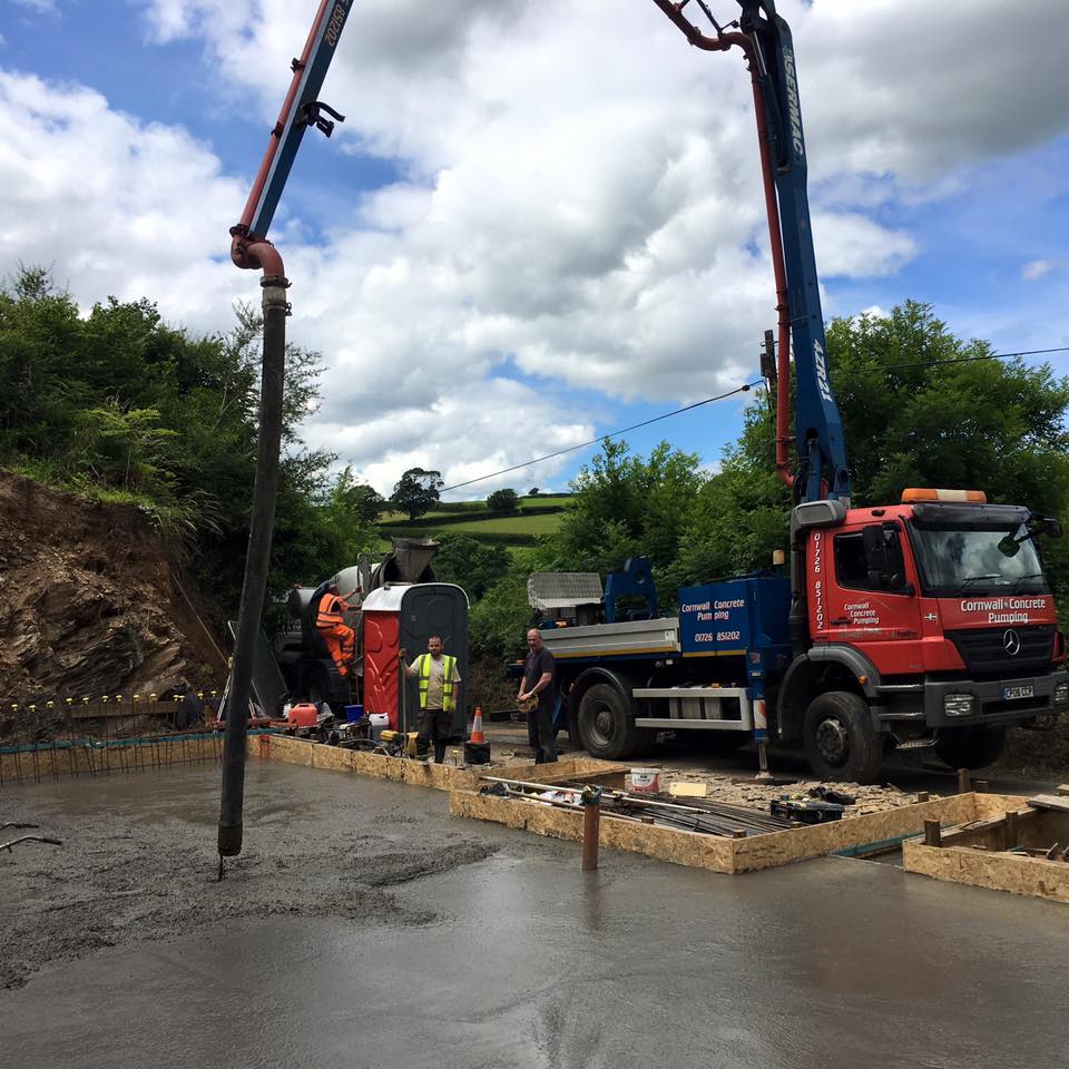 Builders operate a concrete pump truck, pouring wet concrete into wooden forms at a construction site surrounded by greenery and trees—preparing foundations for future kitchen installations or new bathrooms under a partly cloudy sky.