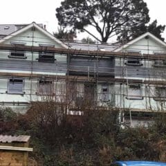 A two-story house with scaffolding around it, indicating property refurbishments in progress. Set on a sloped, overgrown area with trees and bushes in the foreground, a large tree stands behind the building.