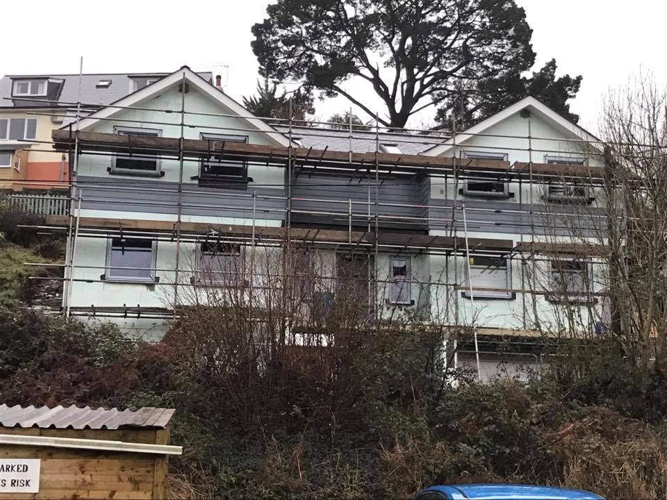 A two-story house with scaffolding around it, indicating property refurbishments in progress. Set on a sloped, overgrown area with trees and bushes in the foreground, a large tree stands behind the building.
