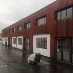 A modern two-story building with wood and white exterior panels, large windows, and a wet parking lot. Known for quality building maintenance, it features a pressure washer near the entrance on a cloudy day.