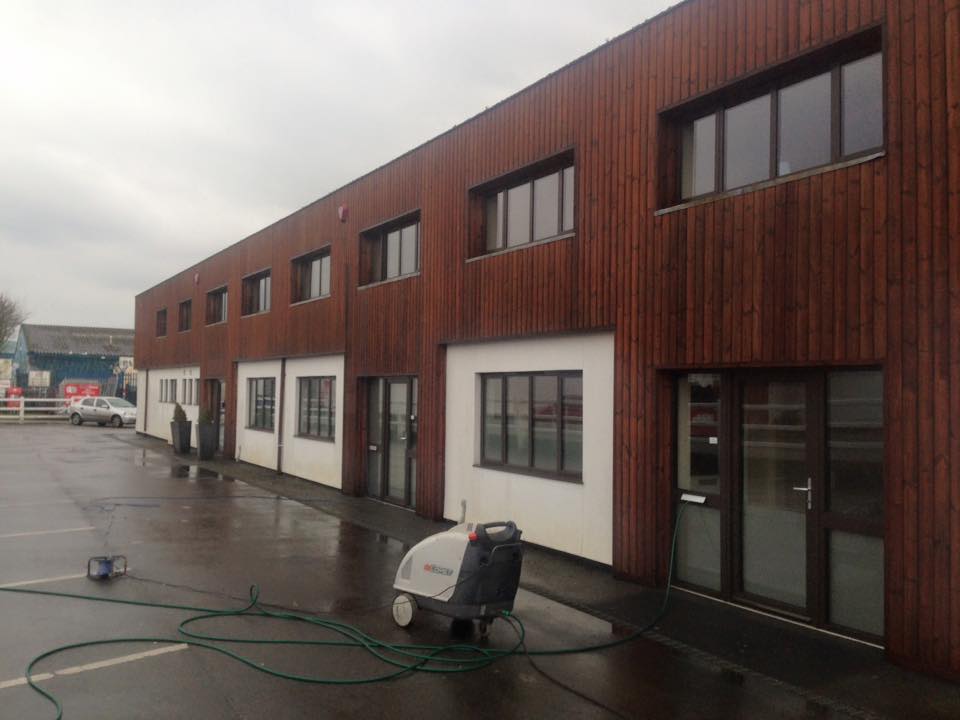 A modern two-story building with wood and white exterior panels, large windows, and a wet parking lot. Known for quality building maintenance, it features a pressure washer near the entrance on a cloudy day.