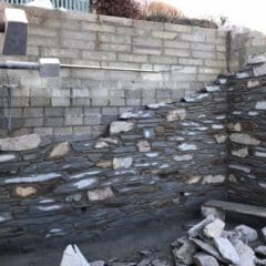 A partially built stone wall stands against an unfinished concrete block wall, with loose stones and construction materials scattered about. Scaffolding to the left hints at ongoing property refurbishments by a skilled builder under a clear sky.