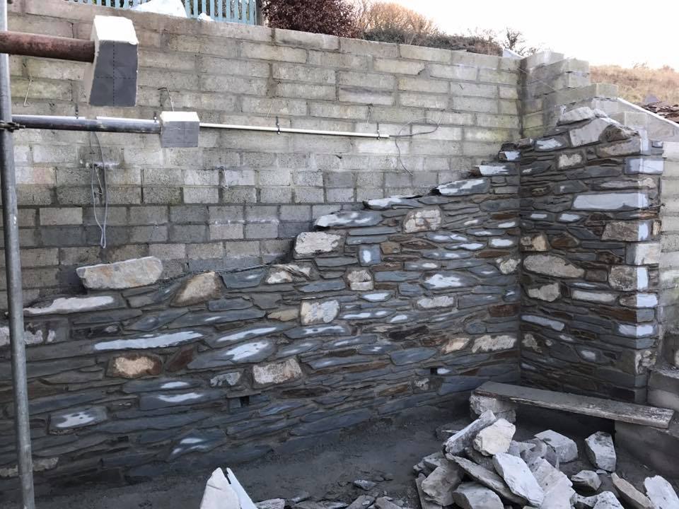 A partially built stone wall stands against an unfinished concrete block wall, with loose stones and construction materials scattered about. Scaffolding to the left hints at ongoing property refurbishments by a skilled builder under a clear sky.