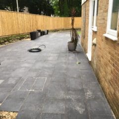 A spacious outdoor patio with large dark gray tiles sits beside a brick house, featuring a tall potted plant and several empty black planters. Perfect for relaxing after property refurbishments, with a coiled hose and wooden fence enclosing the yard.