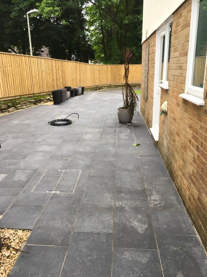 A spacious outdoor patio with large dark gray tiles sits beside a brick house, featuring a tall potted plant and several empty black planters. Perfect for relaxing after property refurbishments, with a coiled hose and wooden fence enclosing the yard.