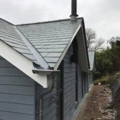 A modern house with blue-grey siding and a slate roof, featuring a prominent chimney and metal gutters—an excellent example of quality building maintenance. The house is surrounded by a dirt path and some trees under a cloudy sky.