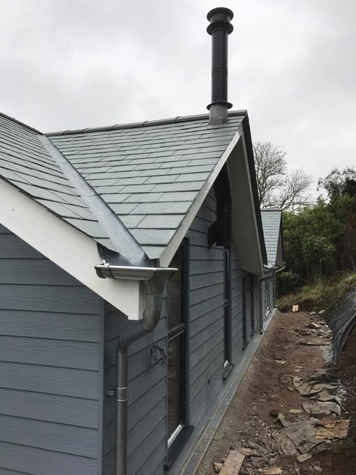 A modern house with blue-grey siding and a slate roof, featuring a prominent chimney and metal gutters—an excellent example of quality building maintenance. The house is surrounded by a dirt path and some trees under a cloudy sky.