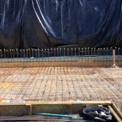 A construction site with a grid of metal rebar on the ground, capped vertical rods, and a black tarp covering the background wall. Builder tools, construction materials, and a wheelbarrow are also visible—perfect for property refurbishments.