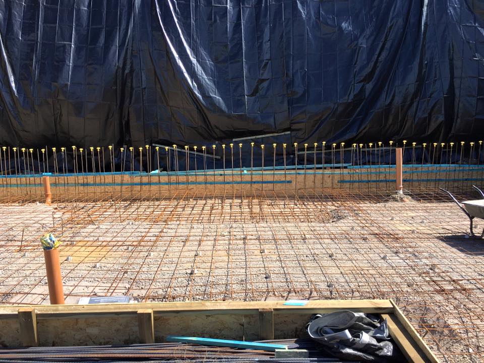 A construction site with a grid of metal rebar on the ground, capped vertical rods, and a black tarp covering the background wall. Builder tools, construction materials, and a wheelbarrow are also visible—perfect for property refurbishments.