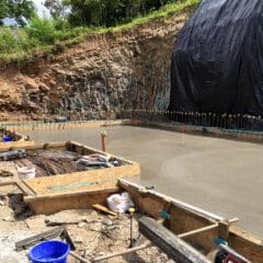 Freshly poured concrete foundation at a construction site, with wooden formwork, construction tools, and materials around—ideal groundwork for future kitchen installations or new bathrooms. The area is bordered by a tall, rocky earth wall partly covered with black tarp.