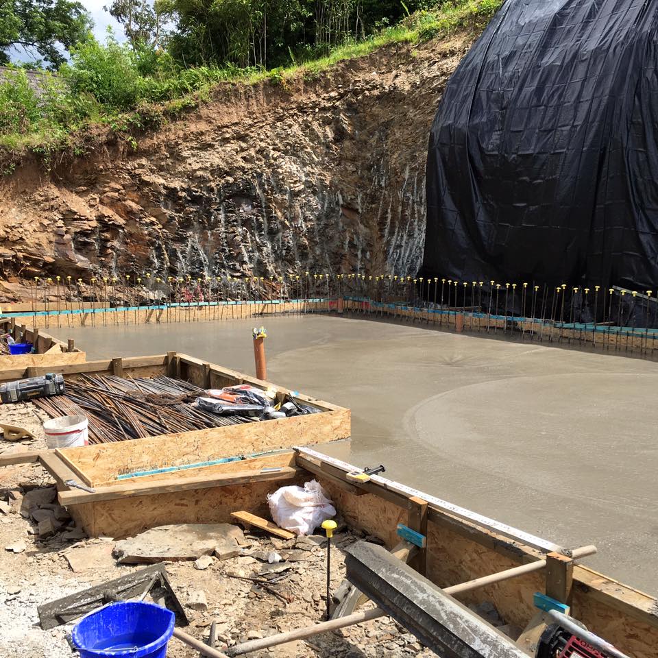 Freshly poured concrete foundation at a construction site, with wooden formwork, construction tools, and materials around—ideal groundwork for future kitchen installations or new bathrooms. The area is bordered by a tall, rocky earth wall partly covered with black tarp.