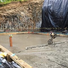 A freshly poured concrete foundation slab is being smoothed with a mechanical tool at a construction site, ready for new bathrooms or kitchen installations. Rebar sticks out from the edges, and plastic sheeting covers the soil wall in the background.