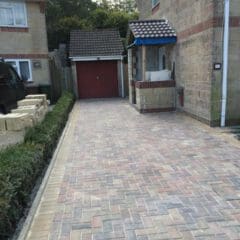 A paved driveway with light and dark rectangular bricks, crafted by an expert builder, leads to a red garage door beside a light-colored brick house with a small porch and hedge-lined border.