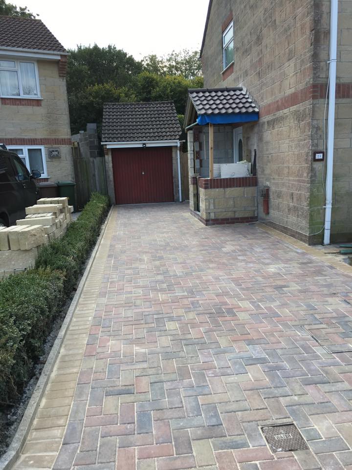 A paved driveway with light and dark rectangular bricks, crafted by an expert builder, leads to a red garage door beside a light-colored brick house with a small porch and hedge-lined border.