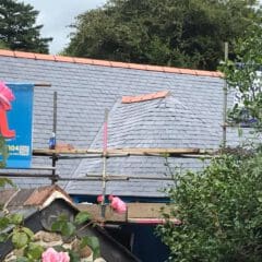 A house roof under construction with grey slate tiles, scaffolding, and a blue banner featuring a parrot. Pink flowers and green foliage are visible in the foreground, highlighting ongoing property refurbishments by an expert builder.