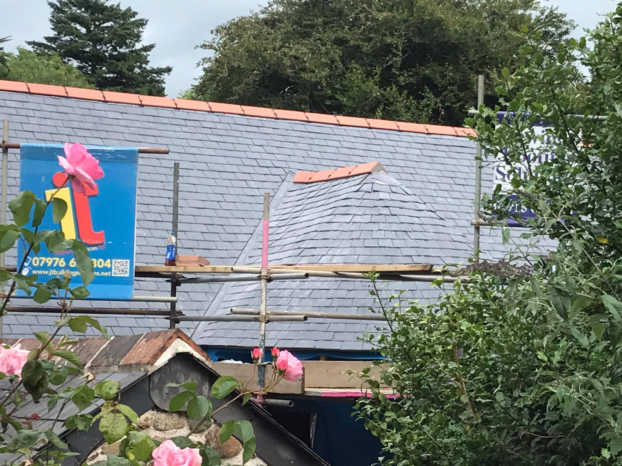 A house roof under construction with grey slate tiles, scaffolding, and a blue banner featuring a parrot. Pink flowers and green foliage are visible in the foreground, highlighting ongoing property refurbishments by an expert builder.