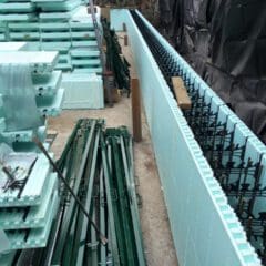 Stacks of light blue insulated concrete forms (ICFs) and green metal braces are arranged at a construction site, where property refurbishments are underway. One ICF wall is partially assembled with visible rebar next to a rock face covered in black sheeting.