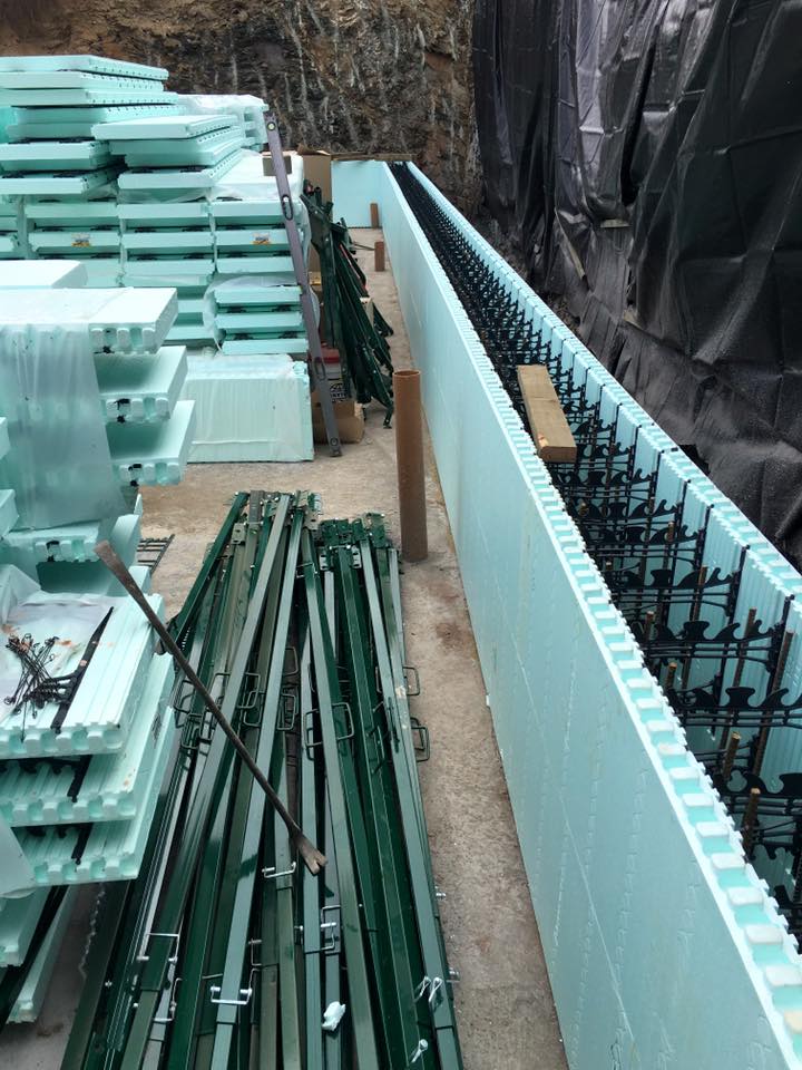 Stacks of light blue insulated concrete forms (ICFs) and green metal braces are arranged at a construction site, where property refurbishments are underway. One ICF wall is partially assembled with visible rebar next to a rock face covered in black sheeting.
