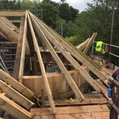 A partially built wooden roof structure with exposed beams and planks, surrounded by scaffolding and tools, set outdoors near trees under a cloudy sky—perfect for inspiring your next property refurbishments or kitchen installations project.