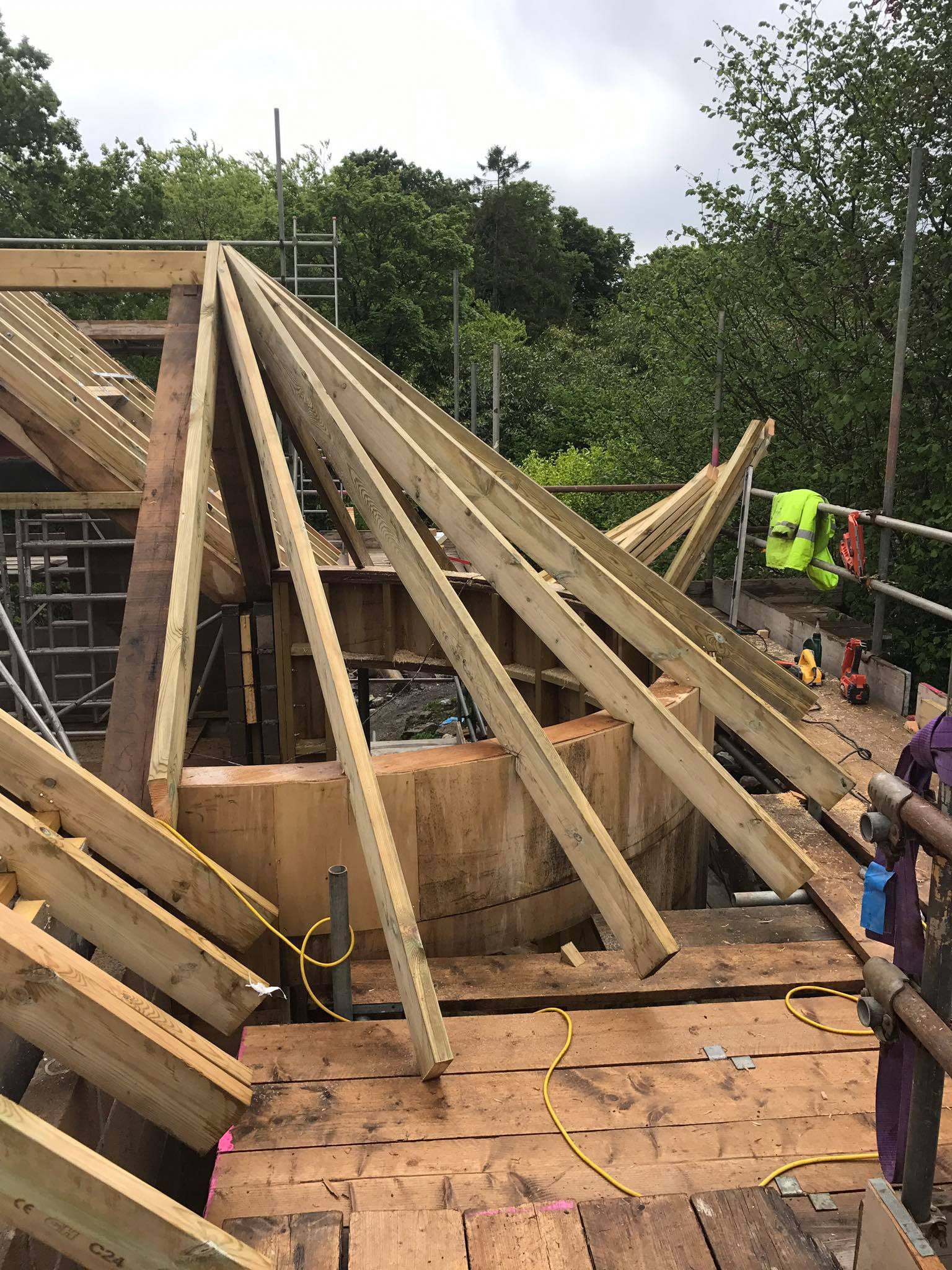 A partially built wooden roof structure with exposed beams and planks, surrounded by scaffolding and tools, set outdoors near trees under a cloudy sky—perfect for inspiring your next property refurbishments or kitchen installations project.