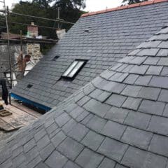 A newly constructed slate roof with a small skylight window and curved section showcases expert builder craftsmanship, surrounded by scaffolding and trees in the background—a perfect example of quality building maintenance.