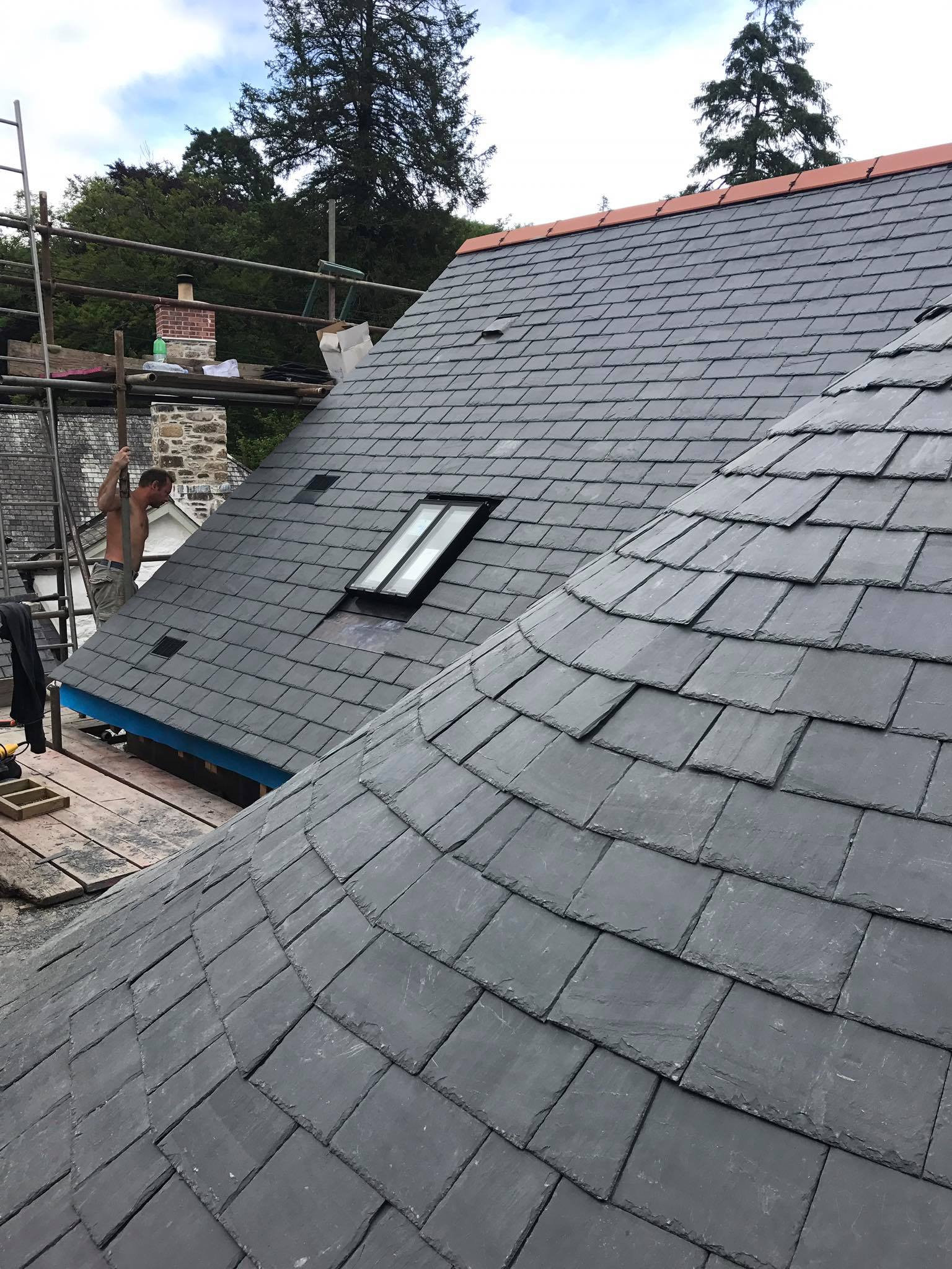 A newly constructed slate roof with a small skylight window and curved section showcases expert builder craftsmanship, surrounded by scaffolding and trees in the background—a perfect example of quality building maintenance.