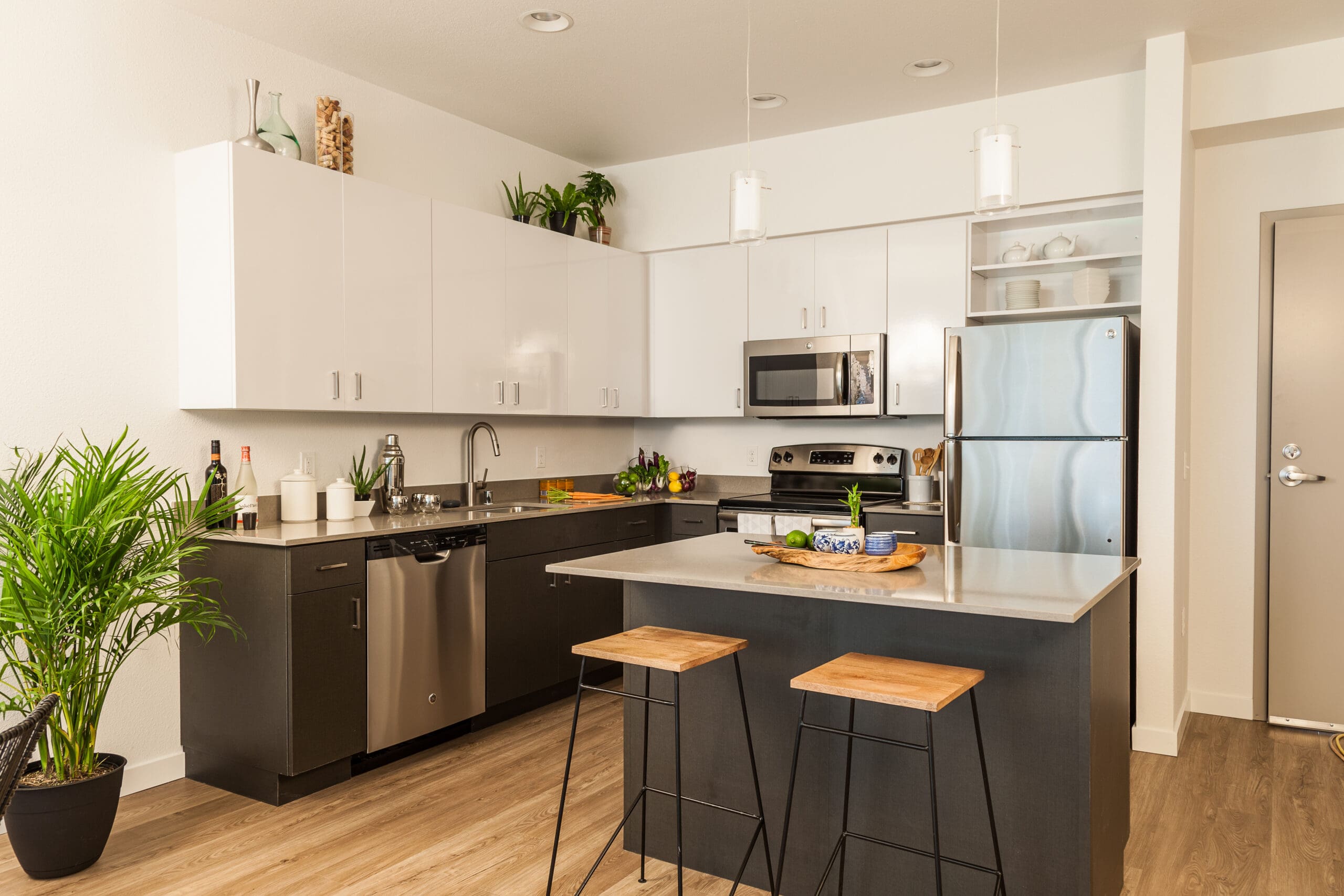 Modern kitchen with white upper cabinets, black lower cabinets, stainless steel appliances, an island with two wooden stools, potted plants, and pendant lights—showcasing quality builder craftsmanship and thoughtful property refurbishments in a bright, organized space.