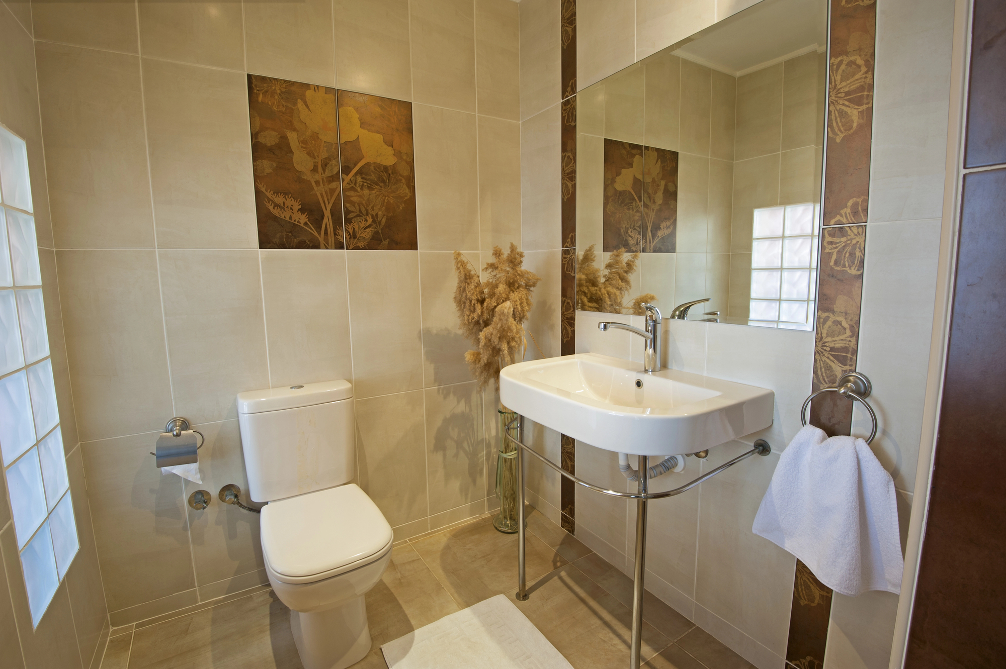 Modern bathroom with a white toilet and sink, large wall mirror, beige tiles with floral patterns, and thoughtful builder touches like a towel ring with a white towel and a glass vase with pampas grass—perfect for stylish property refurbishments.