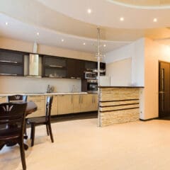 Modern open-plan kitchen and dining area with light wood floors, black and beige cabinetry, a round dining table, stone-accented bar, and recessed ceiling lights—showcasing expert property refurbishments by a skilled builder.